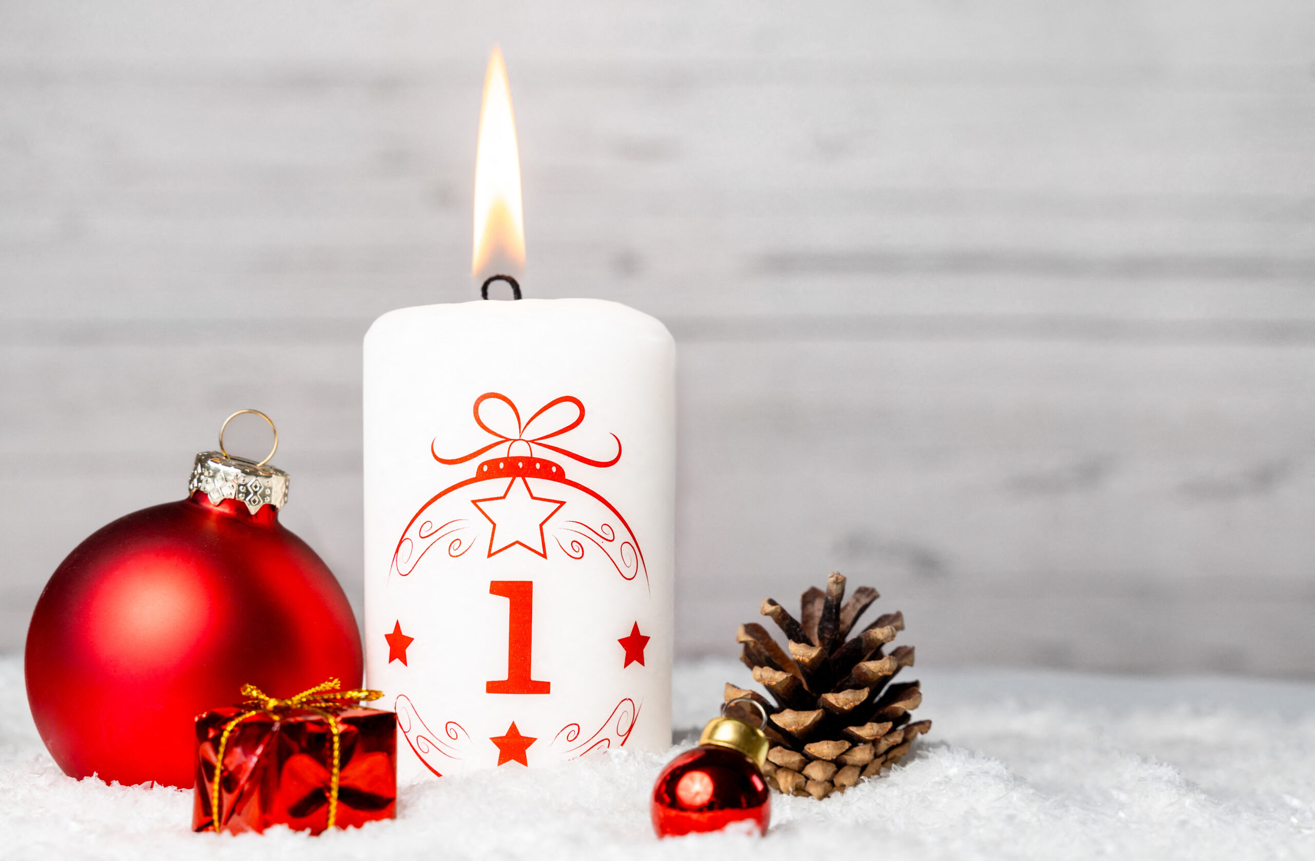 weiße kerze zum 1 advent mit rotem motiv, roten weihnachtskugeln und tannenzapfen im schnee