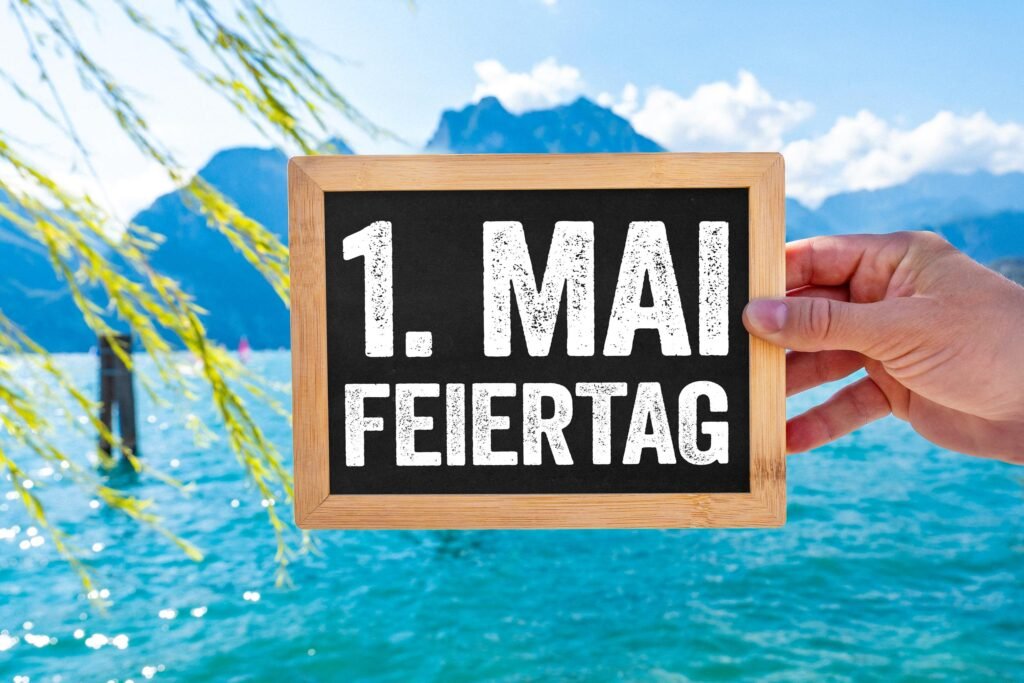 Schild mit Aufschrift 1. Mai Feiertag gehalten vor blauem See und Bergen bei Sonnenschein.