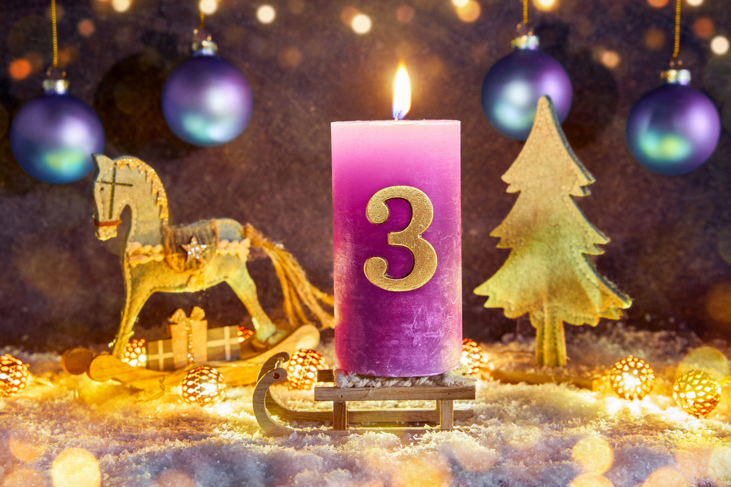 lila kerze mit zahl 3 zum dritten advent, dekoriert mit weihnachtskugeln, tannenzweig und holzpferd im hintergrund
