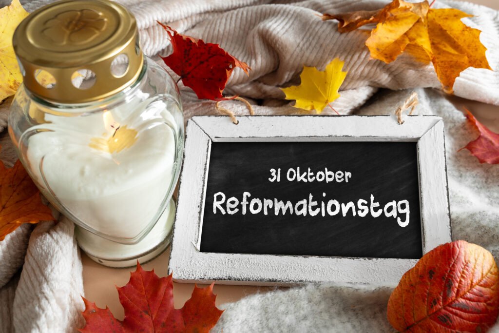 Kerze in Glaslaterne neben Tafel mit der Aufschrift 31. Oktober Reformationstag, umgeben von buntem Herbstlaub auf weicher Decke.
