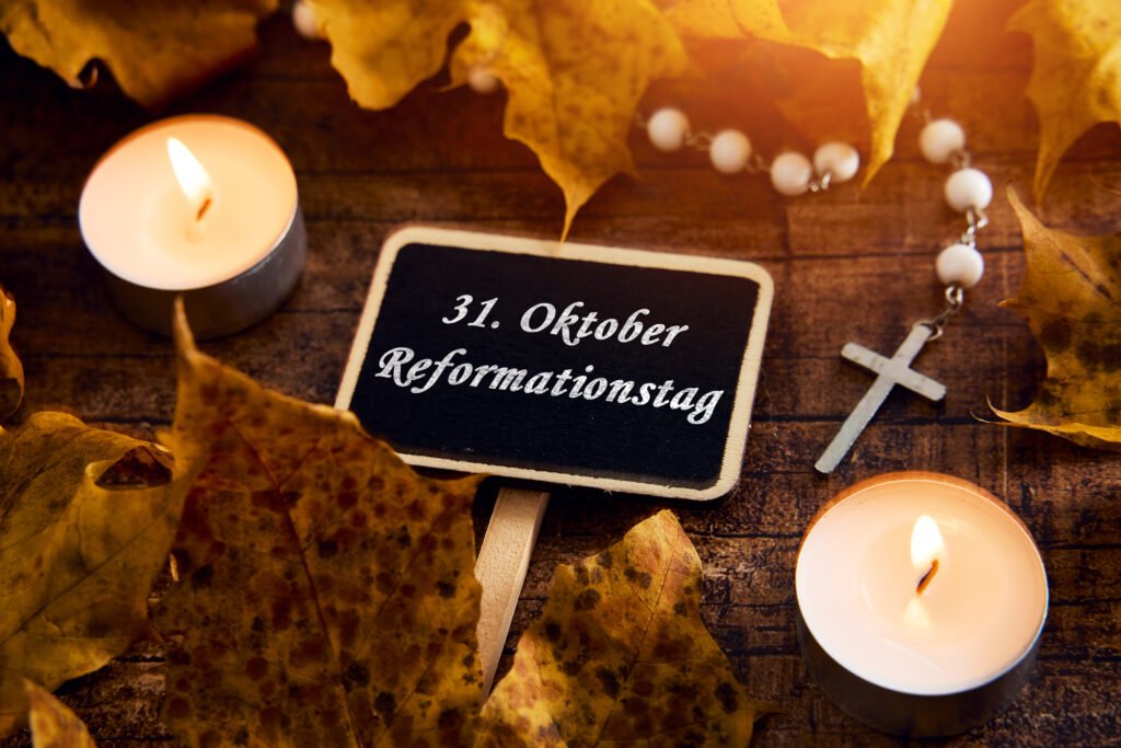 Kleine Tafel mit der Aufschrift 31. Oktober Reformationstag, daneben brennende Kerzen, Kreuz und herbstliche Blätter auf Holztisch.