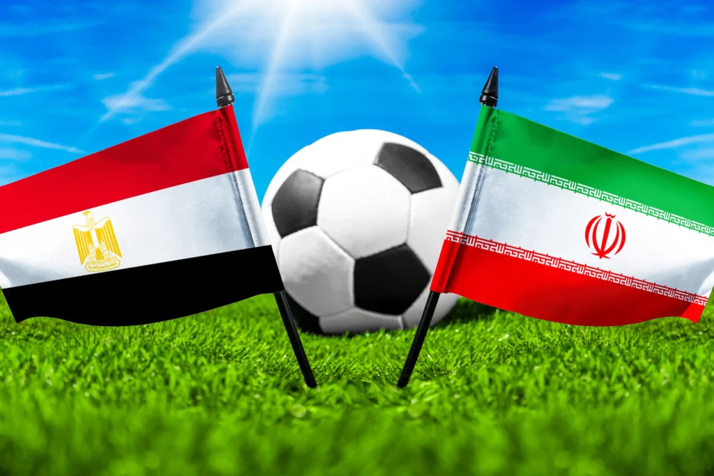 Nationalflaggen von Ägypten und Iran vor einem Fußball auf grünem Rasen unter blauem Himmel.