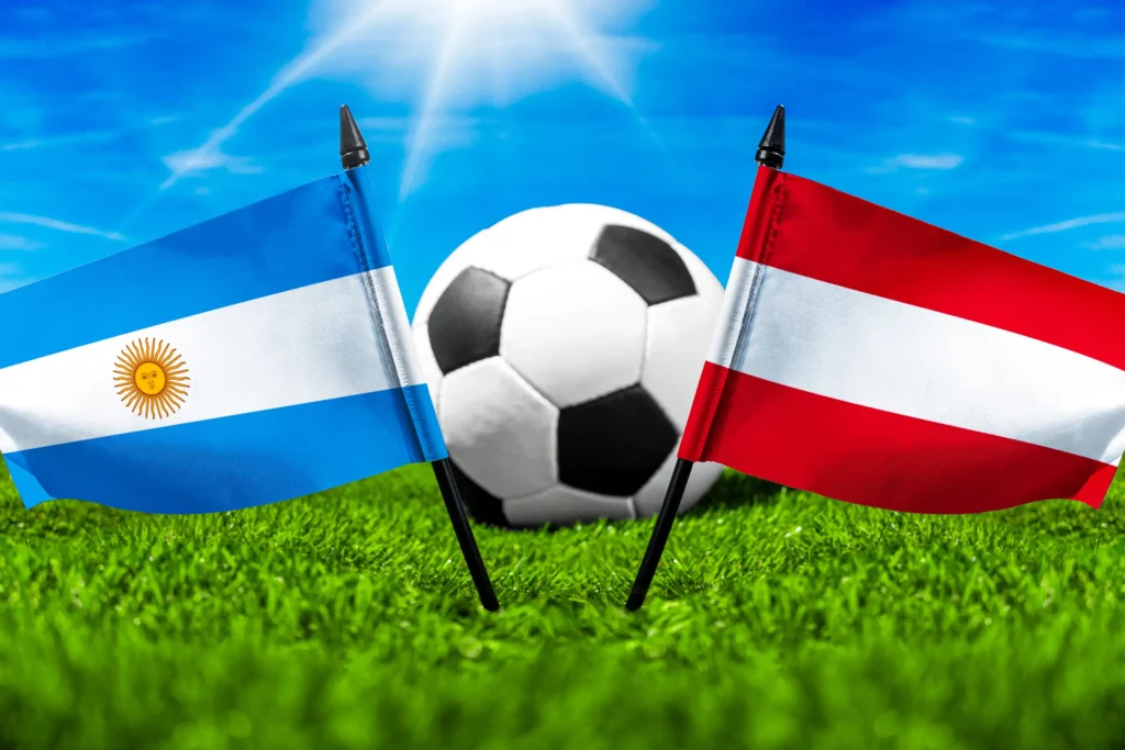 Argentinische und österreichische Flagge vor einem Fußball auf grünem Rasen unter blauem Himmel.