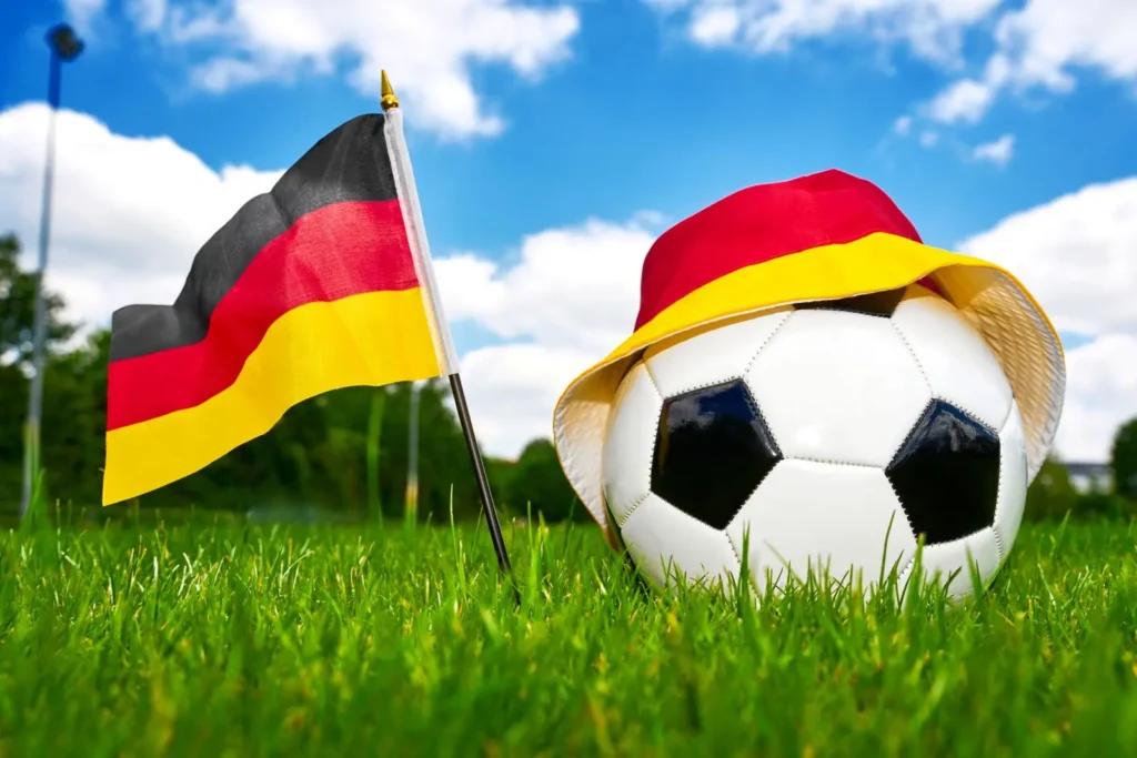 Deutschlandflagge neben Fußball mit schwarz-rot-gelbem Fanhut auf grünem Rasen.