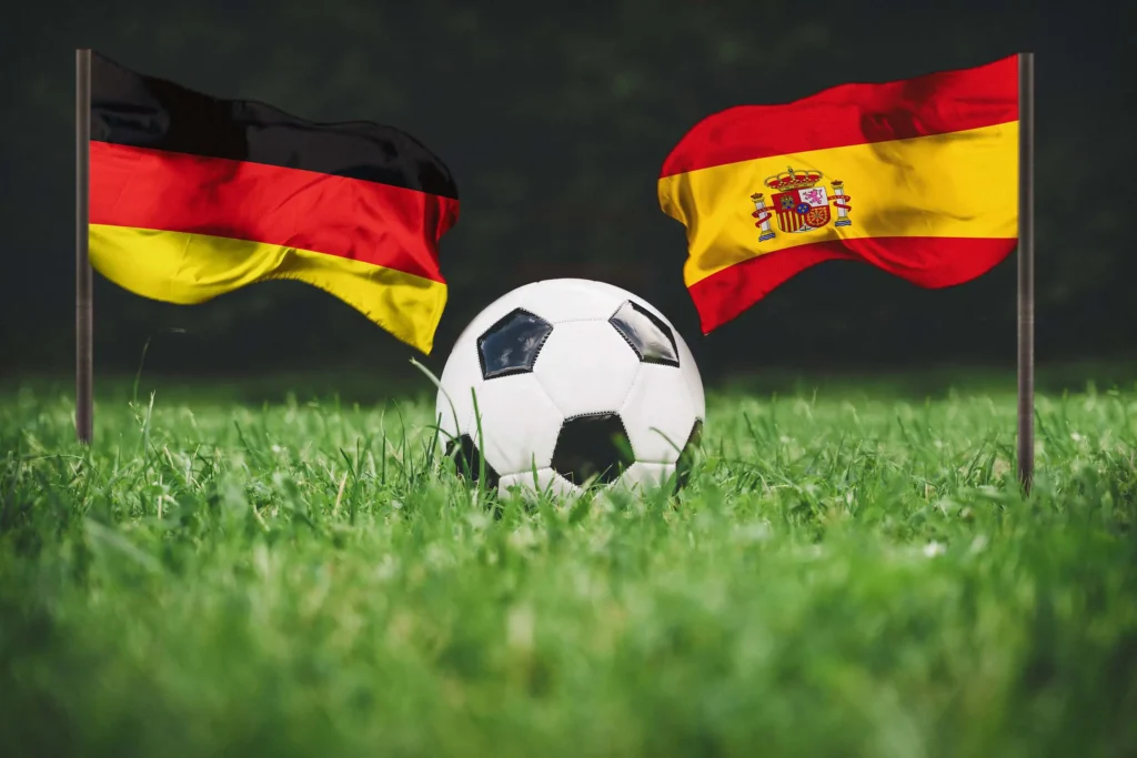 Flaggen von Deutschland und Spanien mit Fußball auf grünem Rasen.