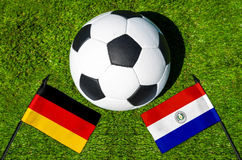 Fußball zwischen den Flaggen von Deutschland und Paraguay auf grünem Rasen als Motiv für ein internationales Fußball-Länderspiel.