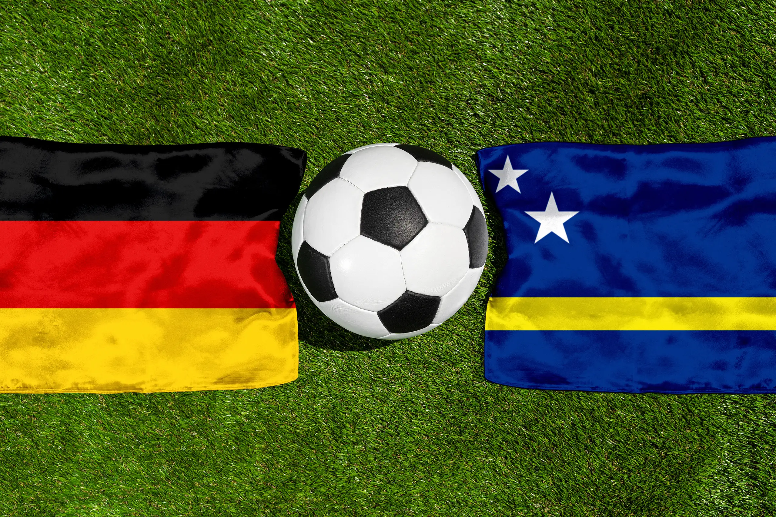 nationalflaggen von deutschland und curaçao neben einem fußball auf grünem spielfeldrasen.