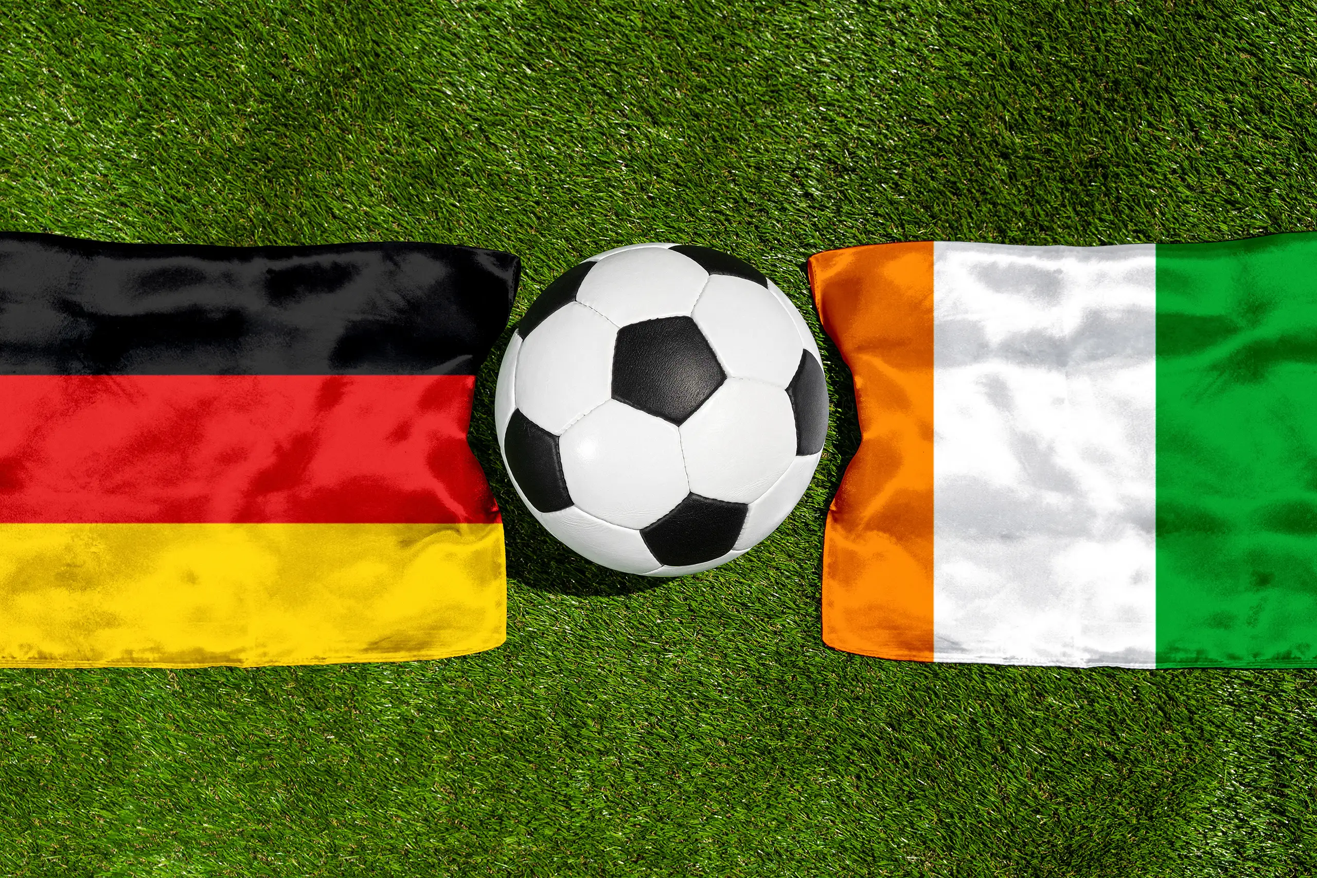 fußball zwischen den flaggen von deutschland und elfenbeinküste auf grünem spielfeldrasen.