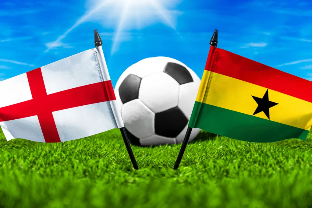 Flaggen von England und Ghana mit Fußball auf grünem Rasen unter blauem Himmel.