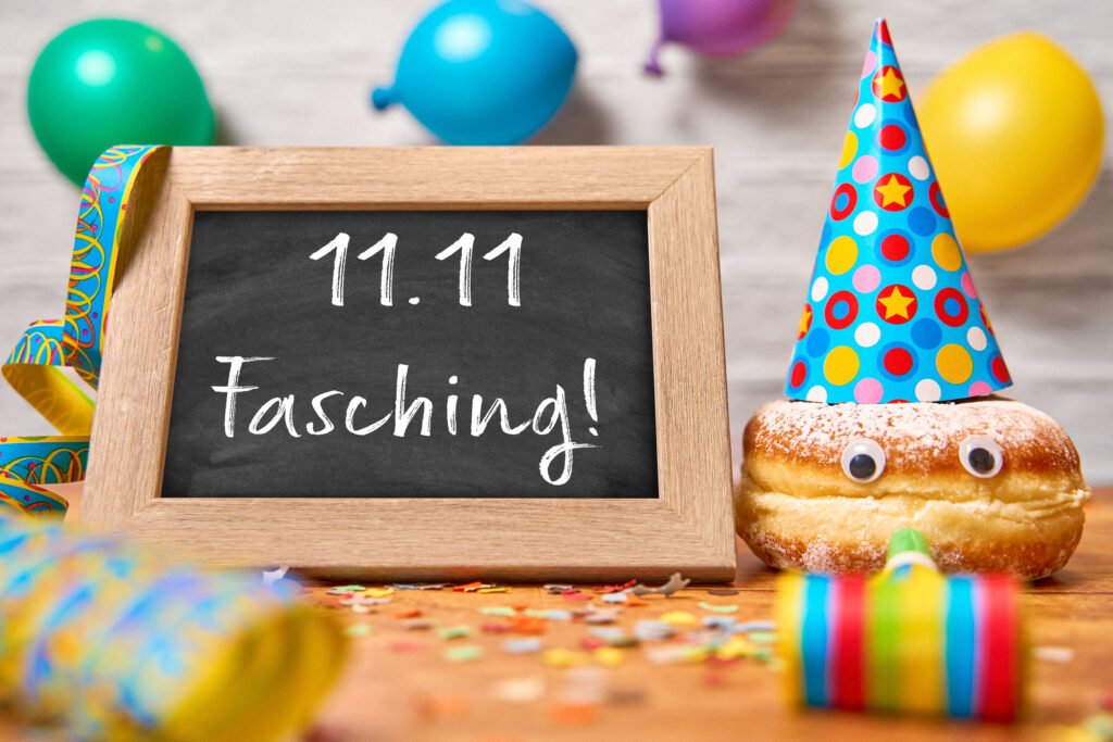Fasching Beginn – Tafel mit Schriftzug „11.11 Fasching!“ und Berliner mit buntem Partyhut, Luftschlangen und Konfetti.
