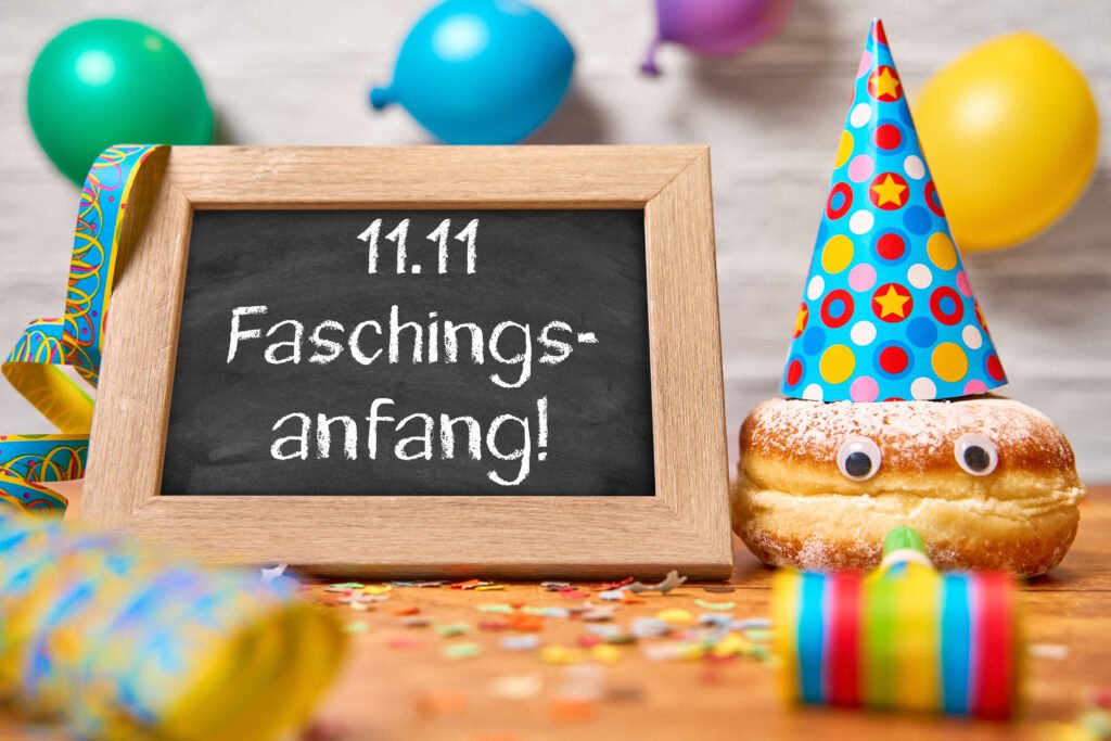 Faschingsanfang am 11. November – Kreidetafel mit Aufschrift „11.11 Faschingsanfang!“ und Berliner mit Partyhut, umgeben von Ballons, Luftschlangen und Konfetti.