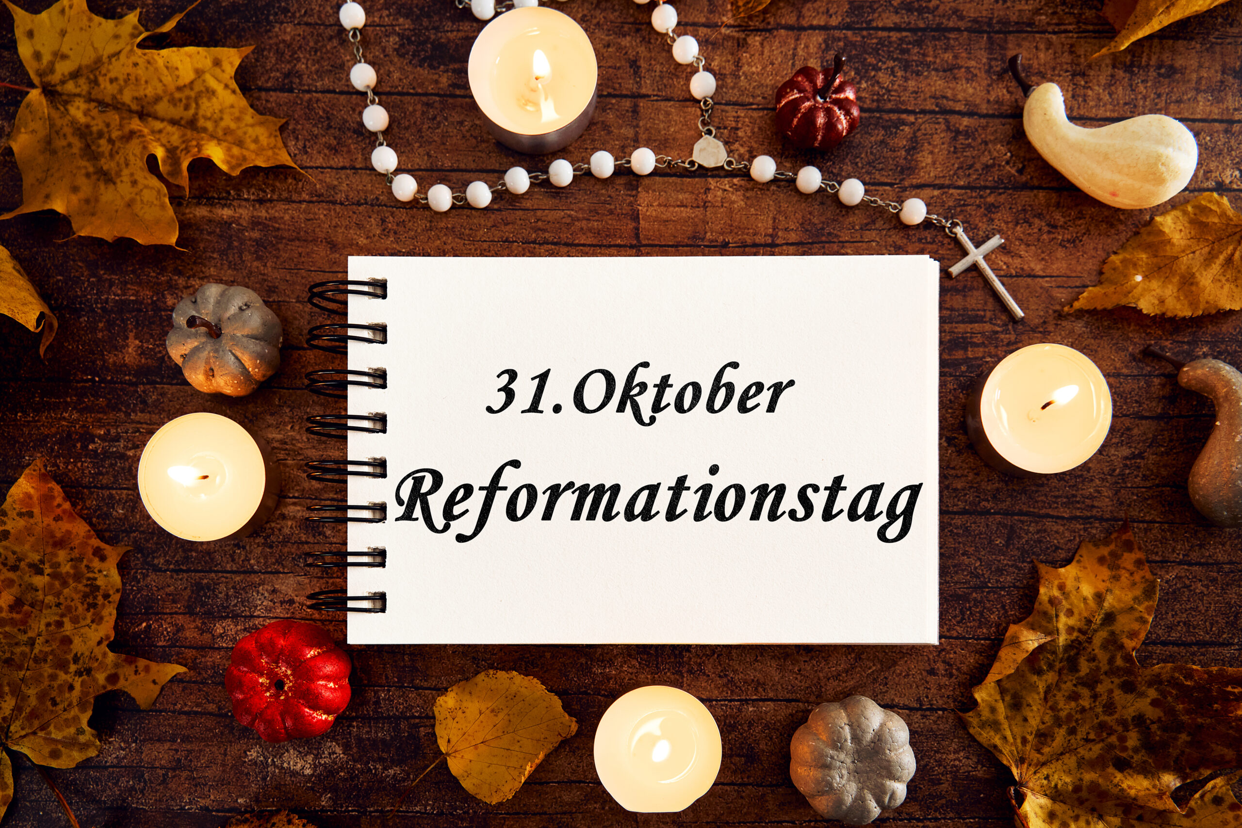 notizbuch mit aufschrift 31. oktober reformationstag, umgeben von brennenden kerzen, kreuz, rosenkranz und herbstlaub auf holztisch.