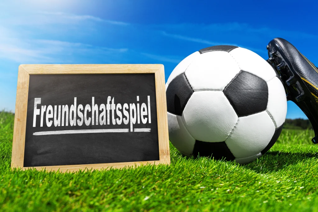 Kreidetafel mit dem Wort Freundschaftsspiel neben Fußball und Fußballschuh auf einem Fußballplatz.