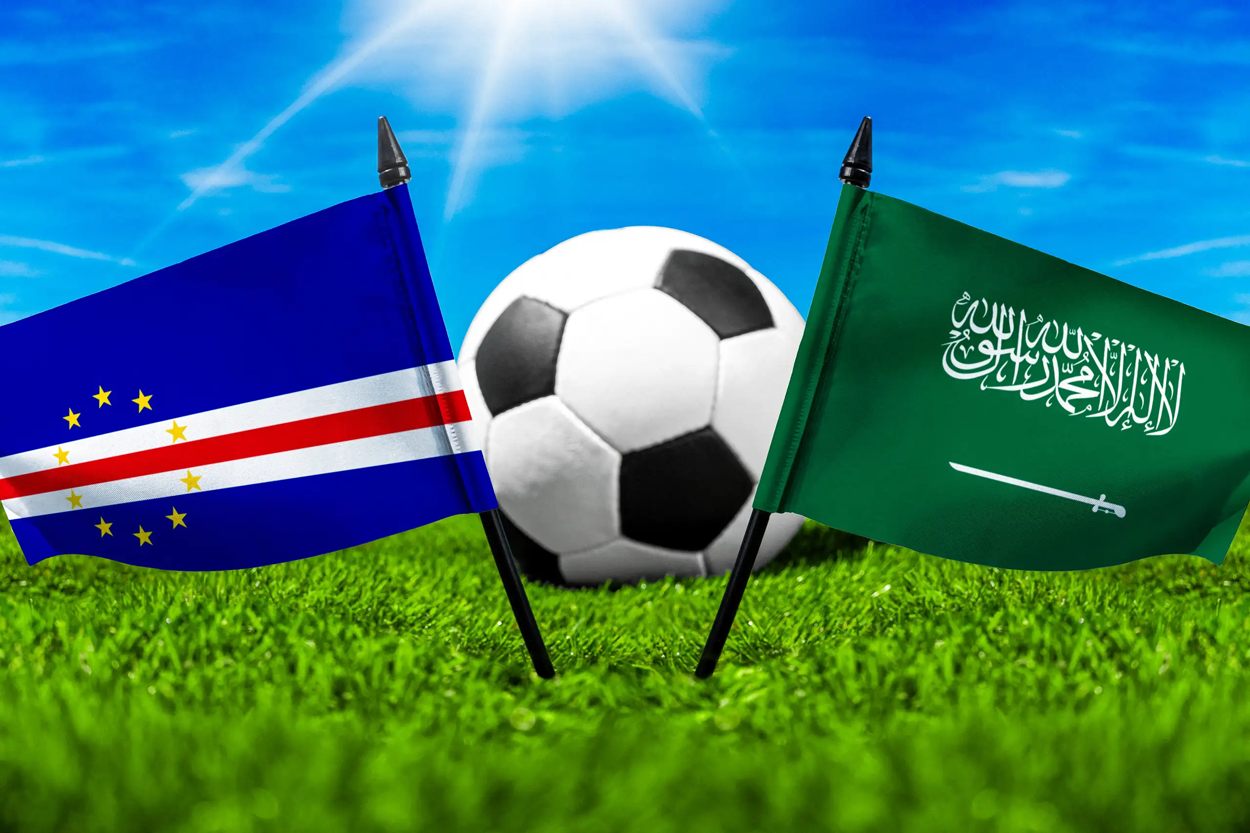 kap verde gegen saudi-arabien mit fußball auf grünem rasen und den nationalflaggen beider länder unter blauem himmel.