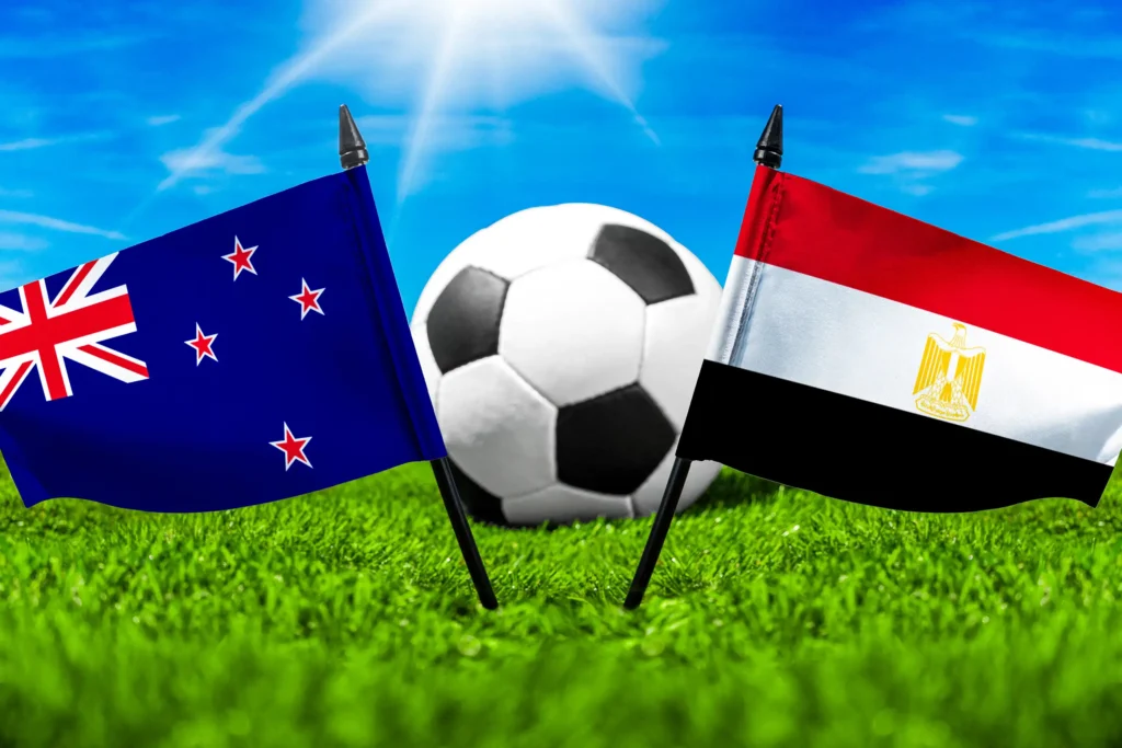 Flaggen von Neuseeland und Ägypten mit Fußball auf grünem Rasen unter blauem Himmel.