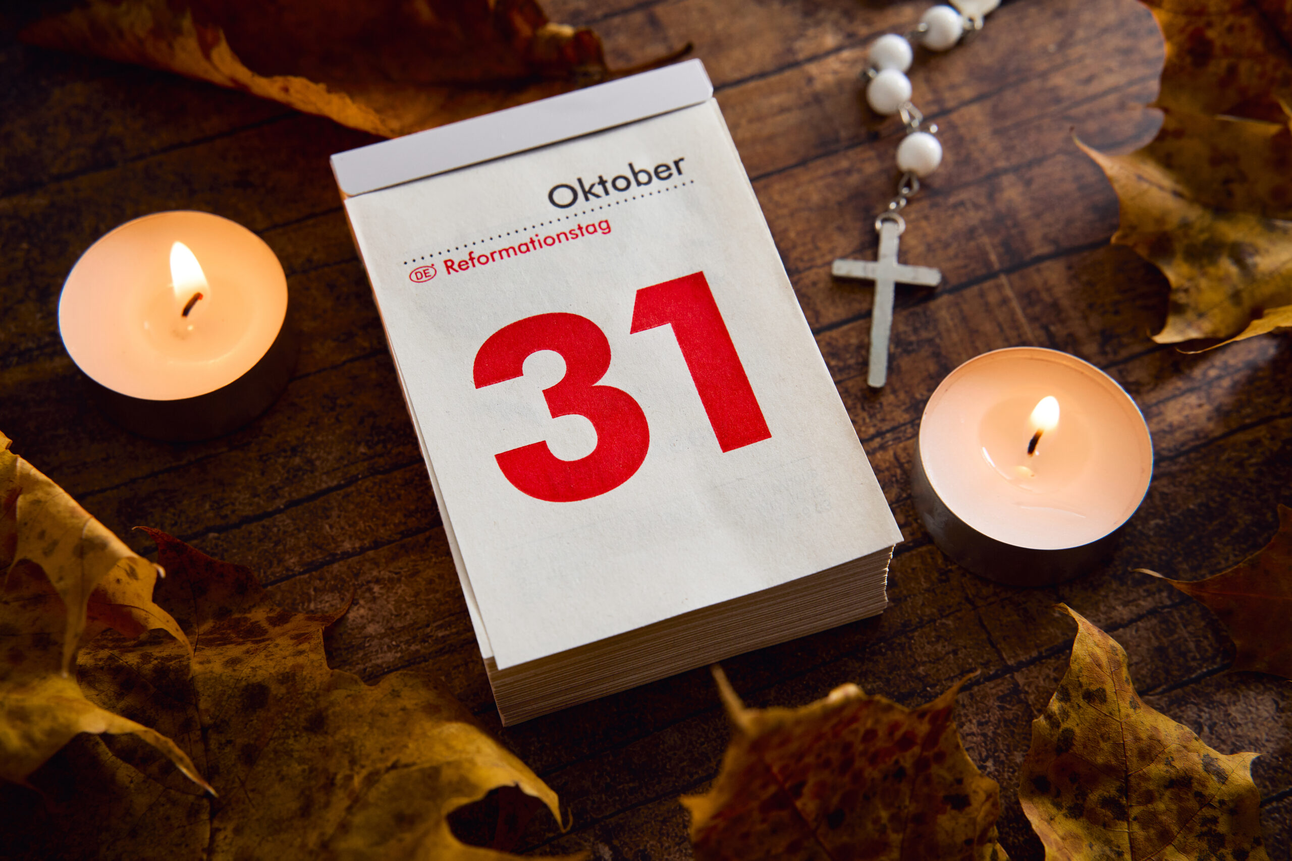 reformationstag 2025 bild mit kalenderblatt vom 31. oktober, brennenden teelichtern, herbstlaub und kreuzkette auf holztisch.