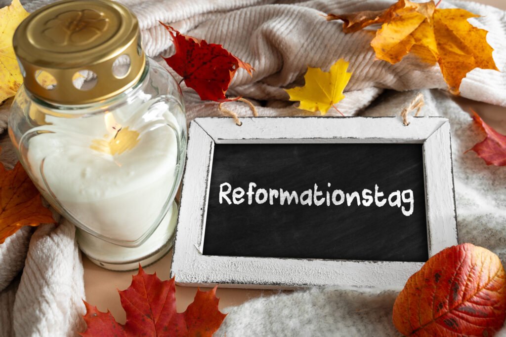 Reformationstag Feiertag Bild mit brennender Kerze in Glaslaterne, Kreidetafel mit Schriftzug „Reformationstag“ und buntem Herbstlaub auf weicher Decke.