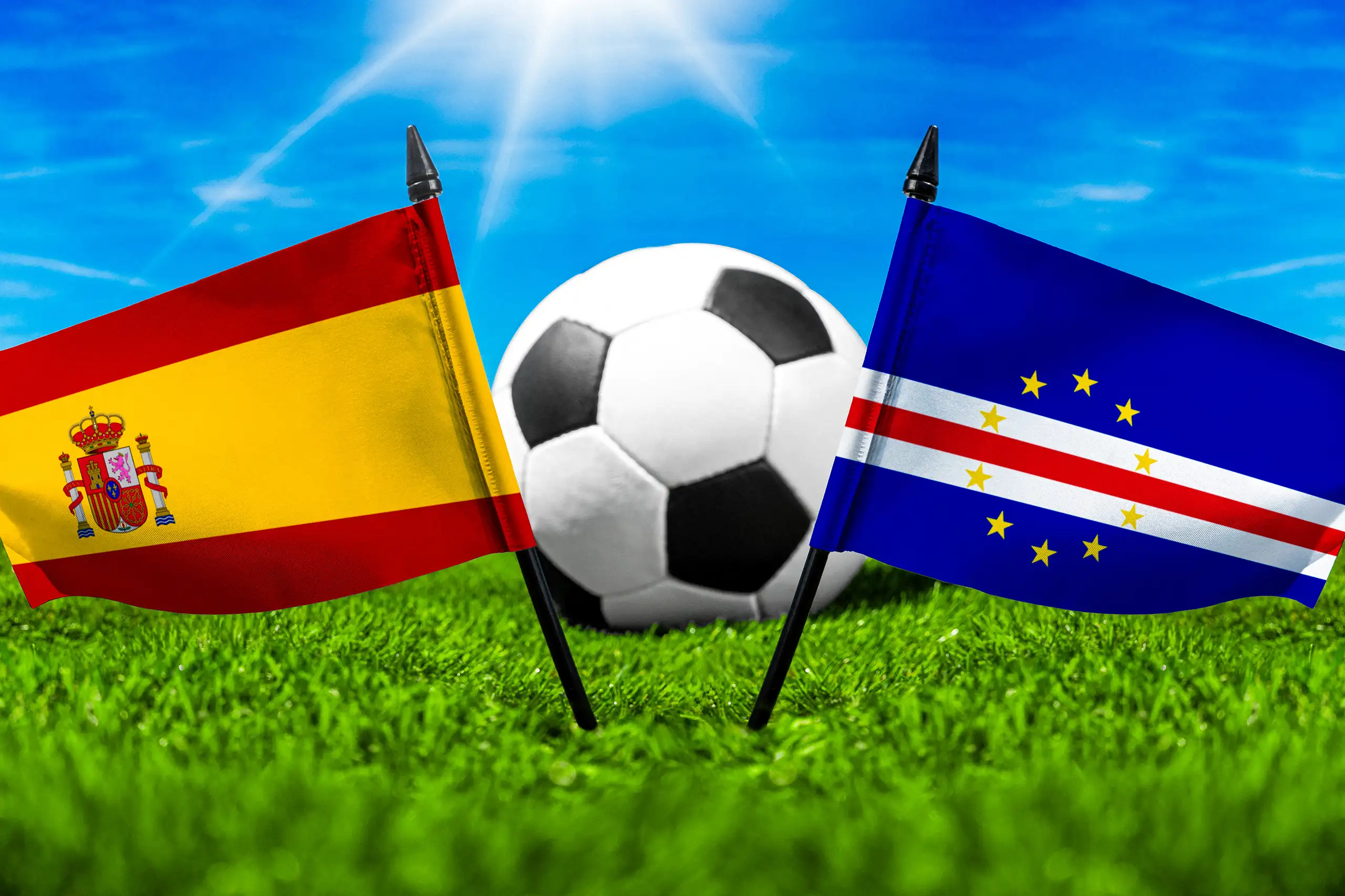 spanische und kapverdische flagge vor einem fußball auf einem spielfeld mit grünem rasen und blauem himmel.