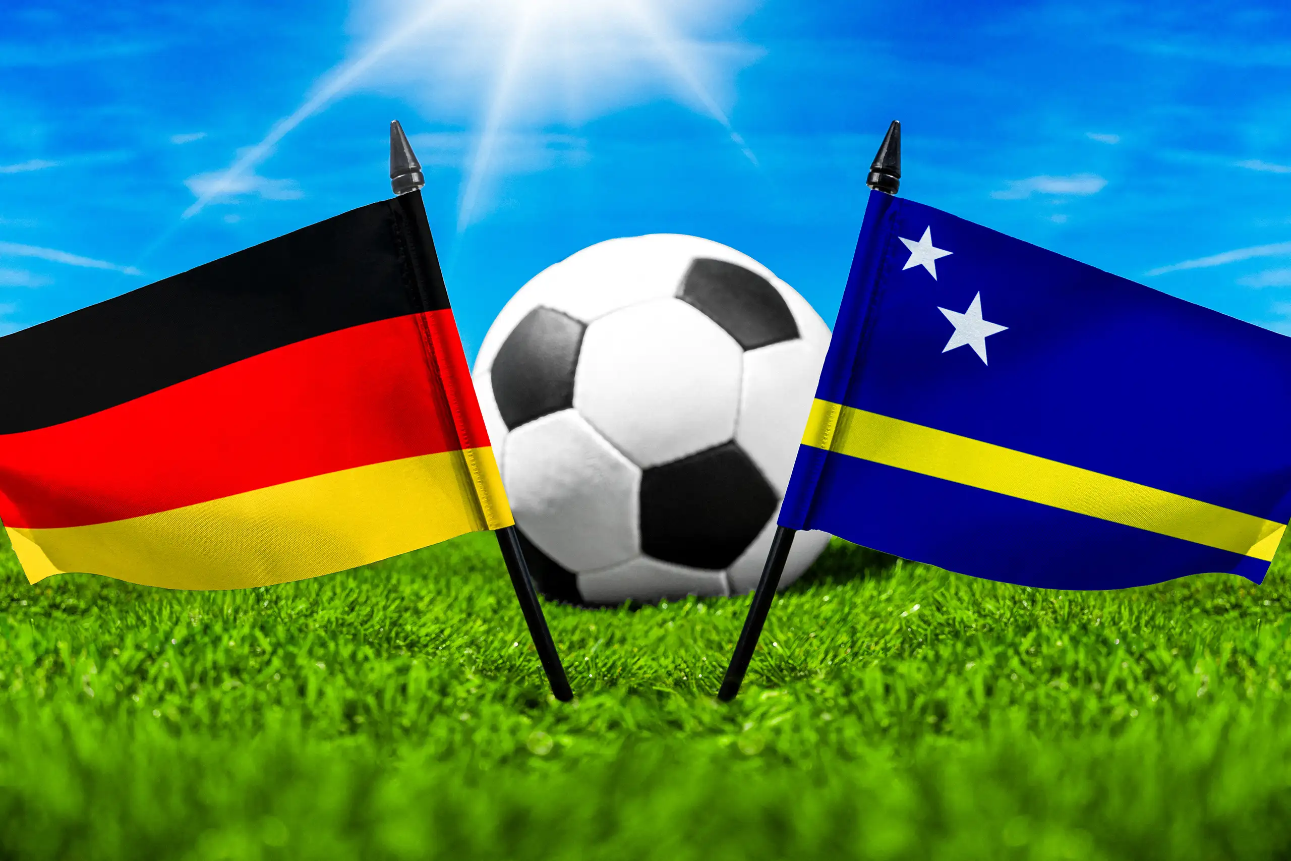flaggen von deutschland und curaçao vor einem fußball auf grünem spielfeldrasen.