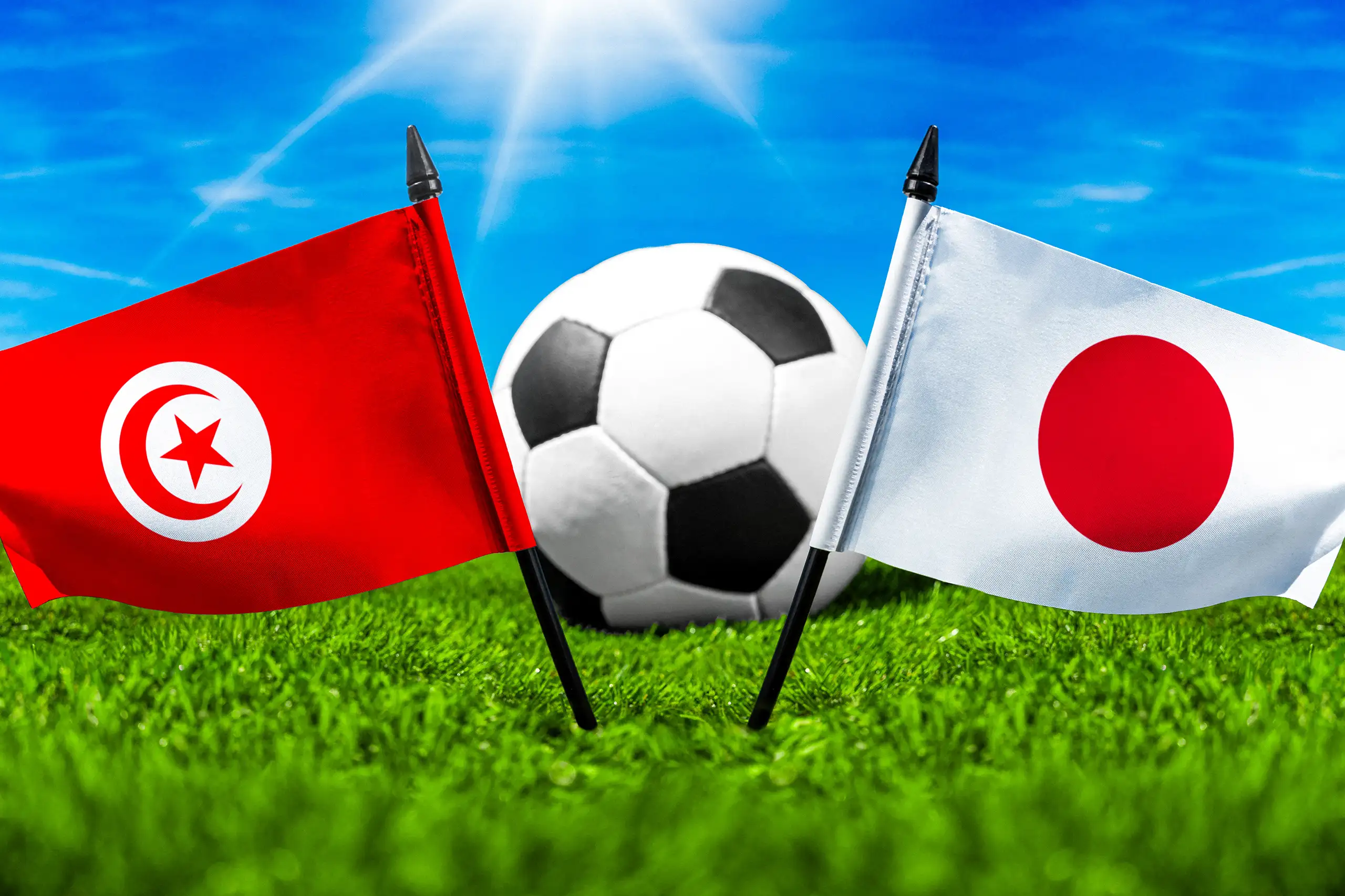 tunesische und japanische nationalflagge mit fußball auf einem rasenplatz unter blauem himmel.