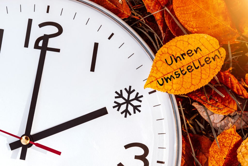 Nahaufnahme einer Uhr mit Herbstblättern und einem Blatt mit der Aufschrift „Uhren umstellen“