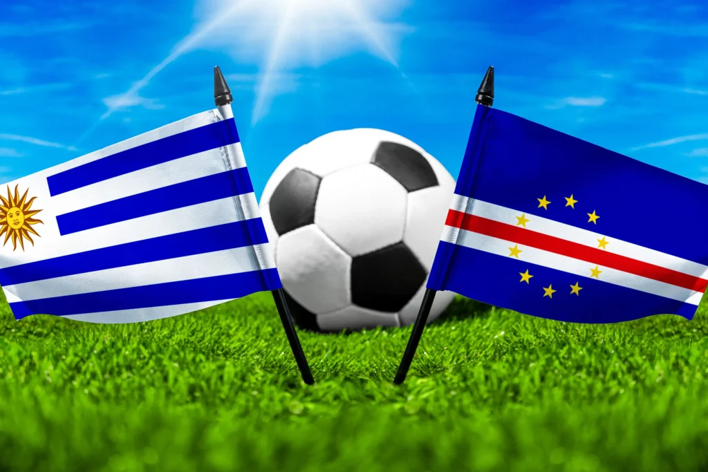 Flaggen von Uruguay und Kap Verde mit Fußball auf grünem Rasen unter blauem Himmel.