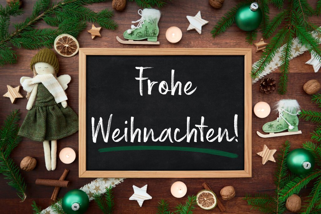 Tafel mit Schrift „Frohe Weihnachten“ umgeben von Weihnachtsdeko, grüne Kugeln, Engel und Tannenzweigen