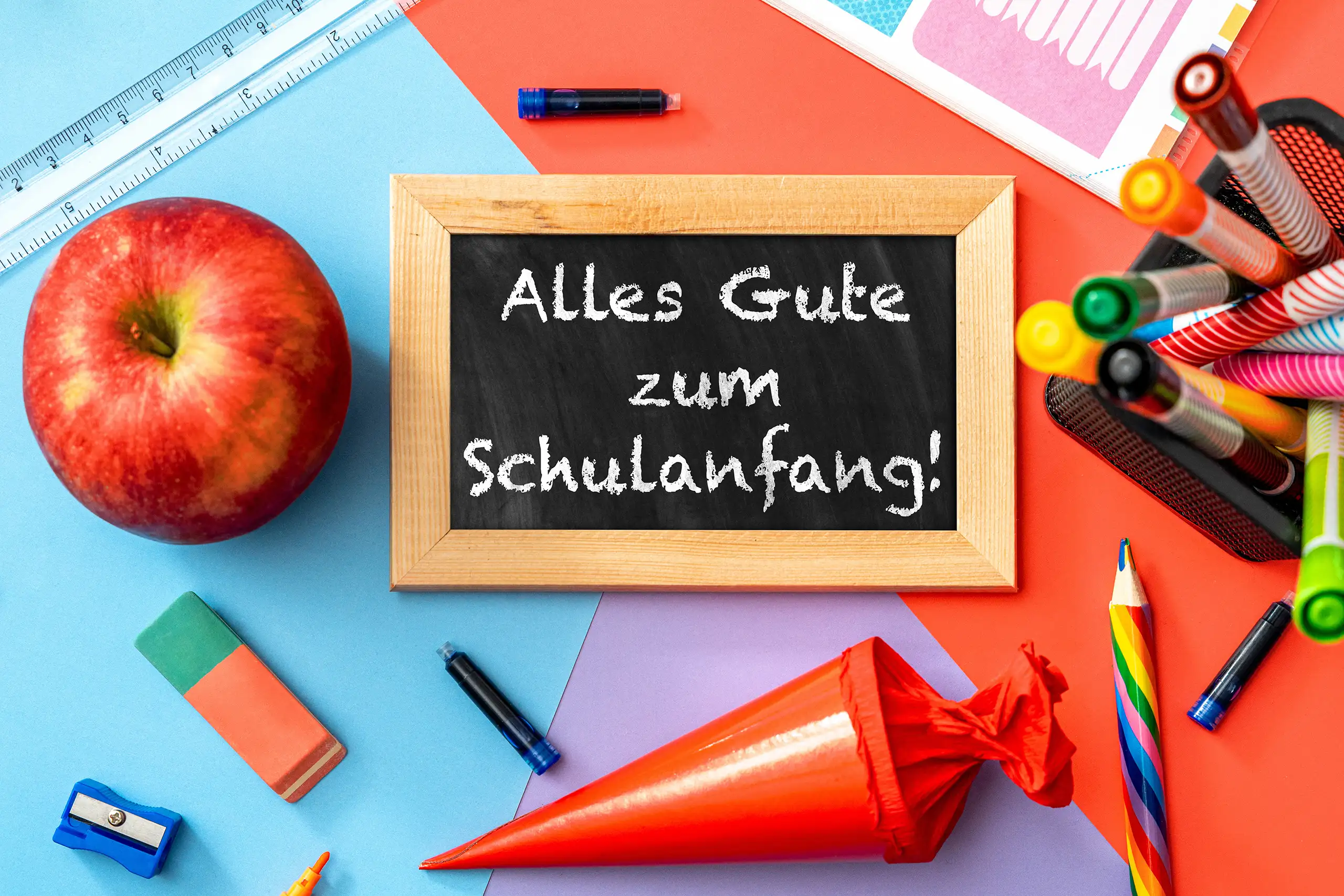 Alles Gute zum Schulanfang auf einer schwarzen Tafel zwischen roter Schultüte, Apfel und bunten Schreibstiften