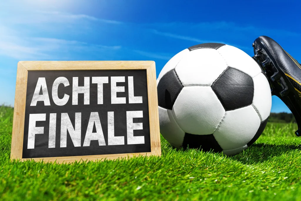 Tafel mit der Aufschrift Achtelfinale neben Fußball und Fußballschuh auf grünem Rasen.
