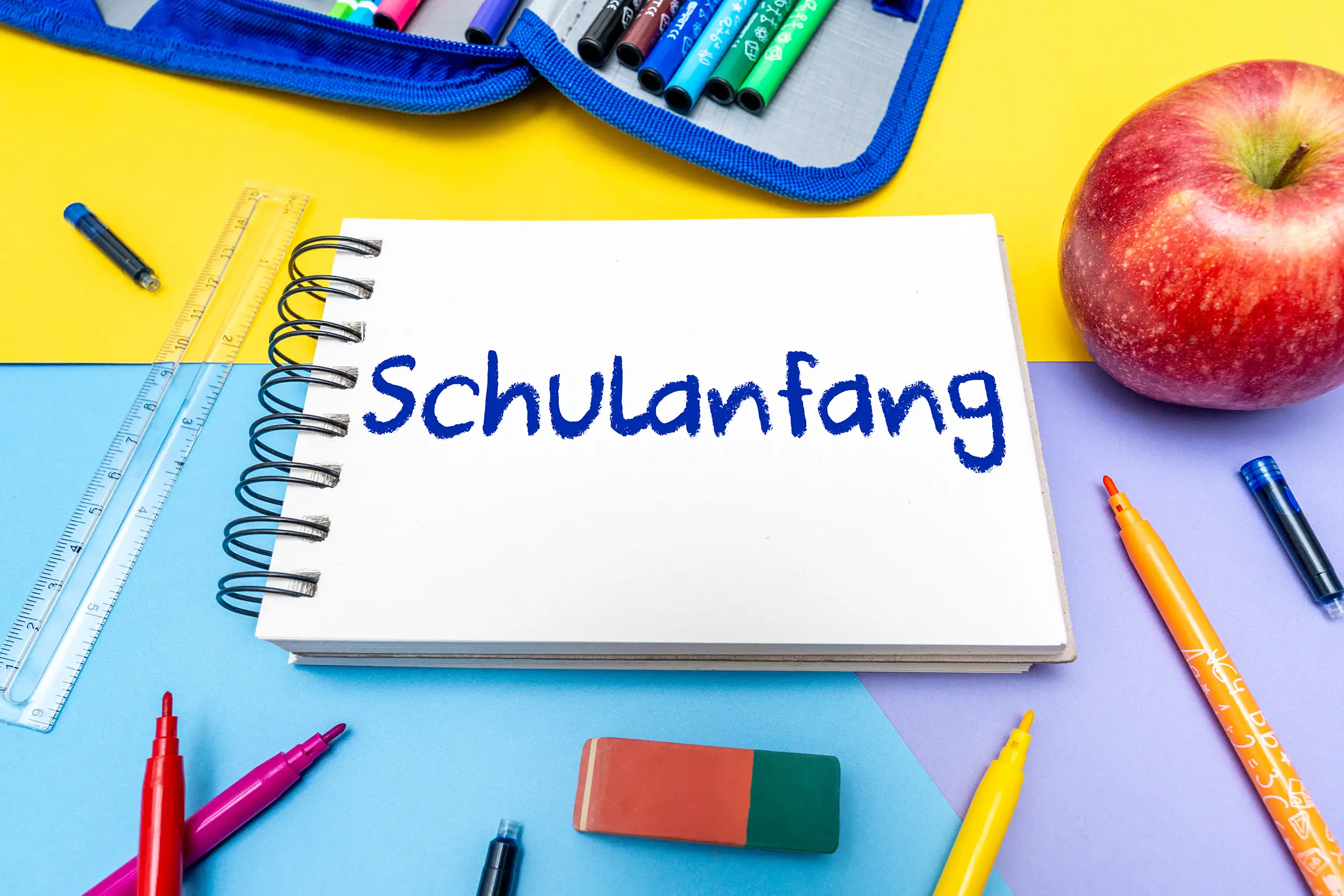 Schulanfang Bild mit beschriftetem Notizbuch, Federmäppchen, bunten Stiften, Linealen und Apfel zum Schulbeginn
