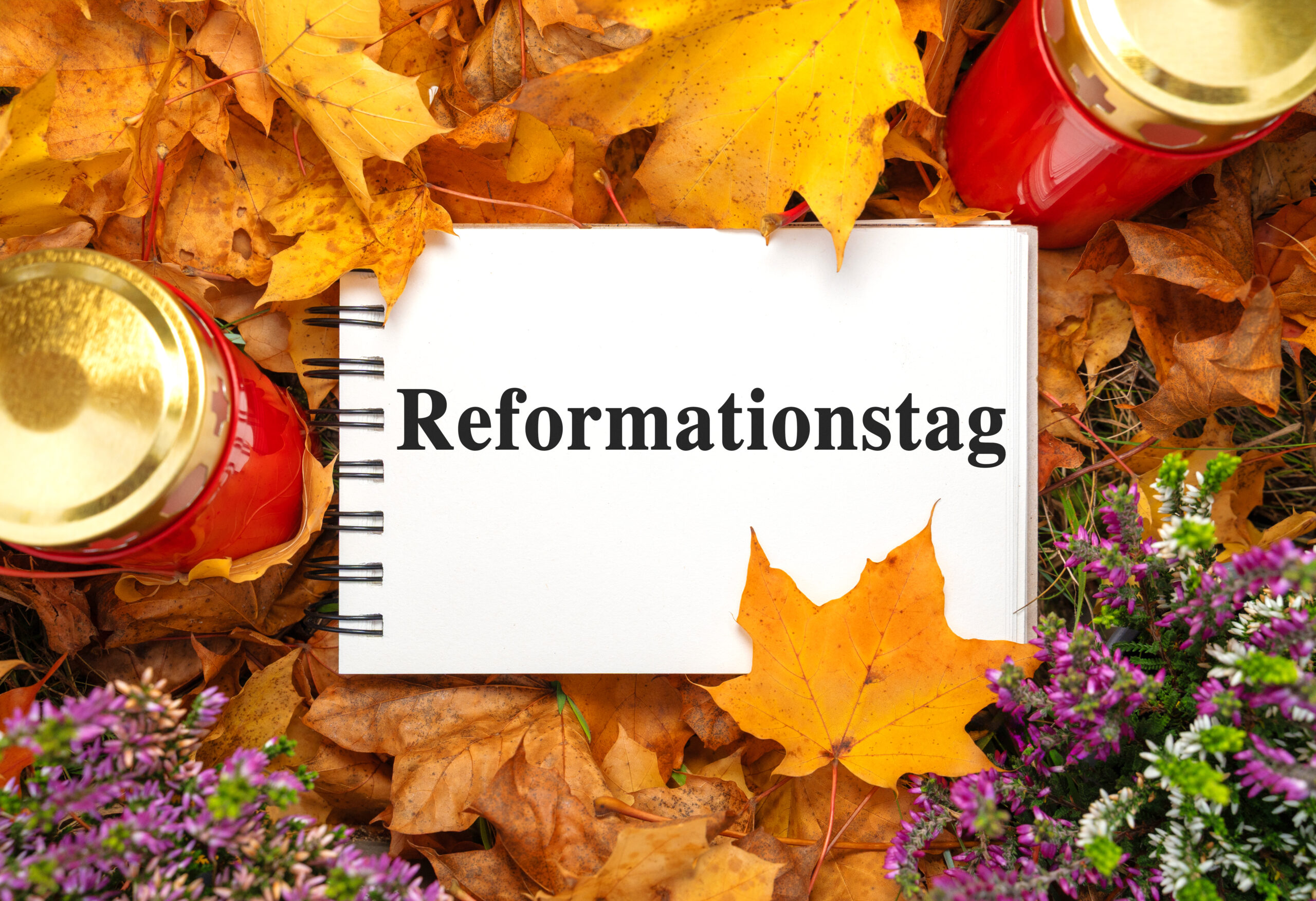notizbuch mit dem wort reformationstag liegt zwischen buntem herbstlaub, roten grablichtern und violetten heideblüten.