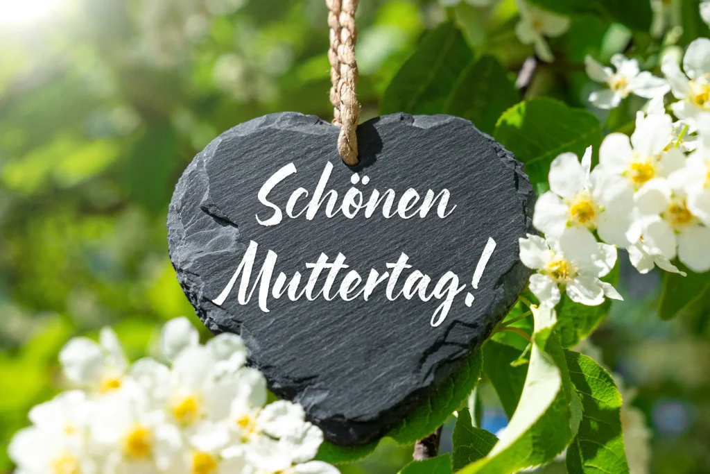 Eleganter Muttertagsgruß mit Schönen Muttertag Schriftzug auf schwarzem Herzschild zwischen weißen Blüten und grünen Blättern.