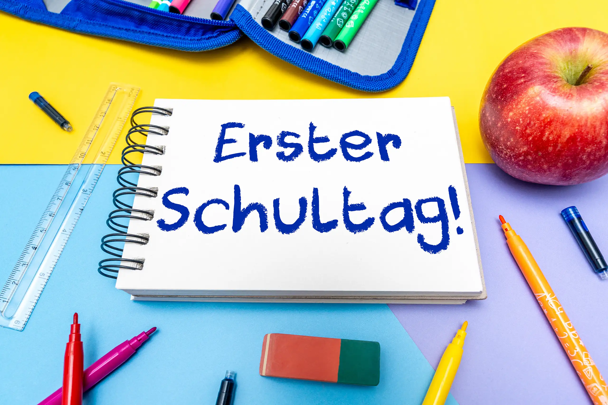 Erster Schultag als blauer Schriftzug auf einem Notizbuch zwischen bunten Stiften, Lineal und Apfel