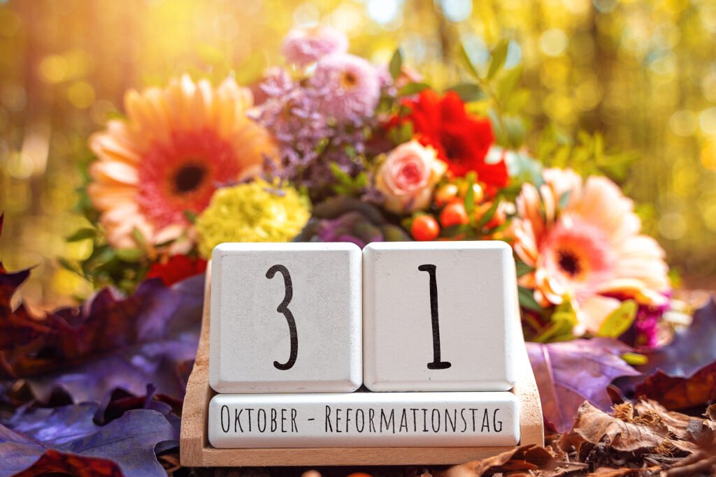 Kalenderblöcke mit dem Datum 31. Oktober Reformationstag, umgeben von bunten Blumen und Herbstlaub im warmen Sonnenlicht.
