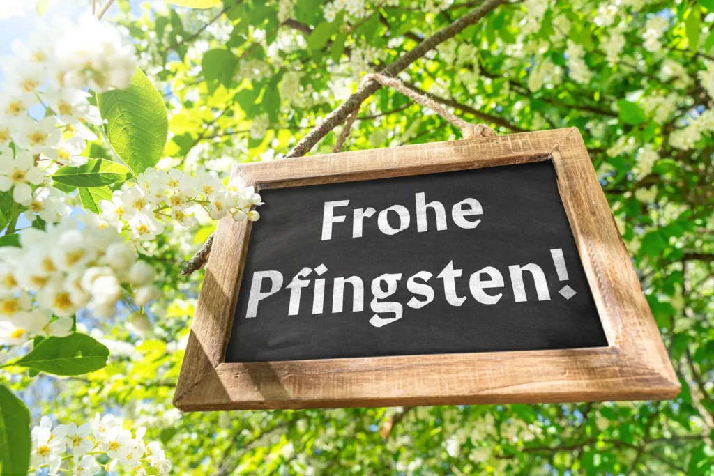 Holztafel mit dem Schriftzug Frohe Pfingsten zwischen weißen Blüten und grünen Blättern an einem Baum im Sonnenlicht.