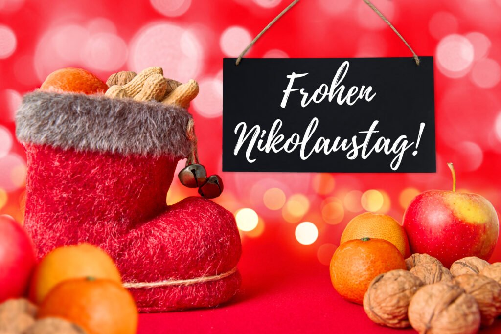 Frohen Nikolaustag – festlicher Nikolausgruß mit rotem Stiefel, Nüssen, Apfel und Mandarinen vor rotem Bokeh-Hintergrund