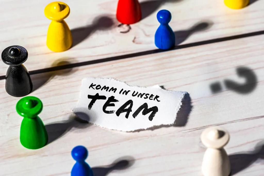 Bunte Spielfiguren auf einer Holzoberfläche umgeben einen Papierzettel mit der Aufschrift Komm in unser Team, daneben ist der Schatten eines Fragezeichens zu sehen.