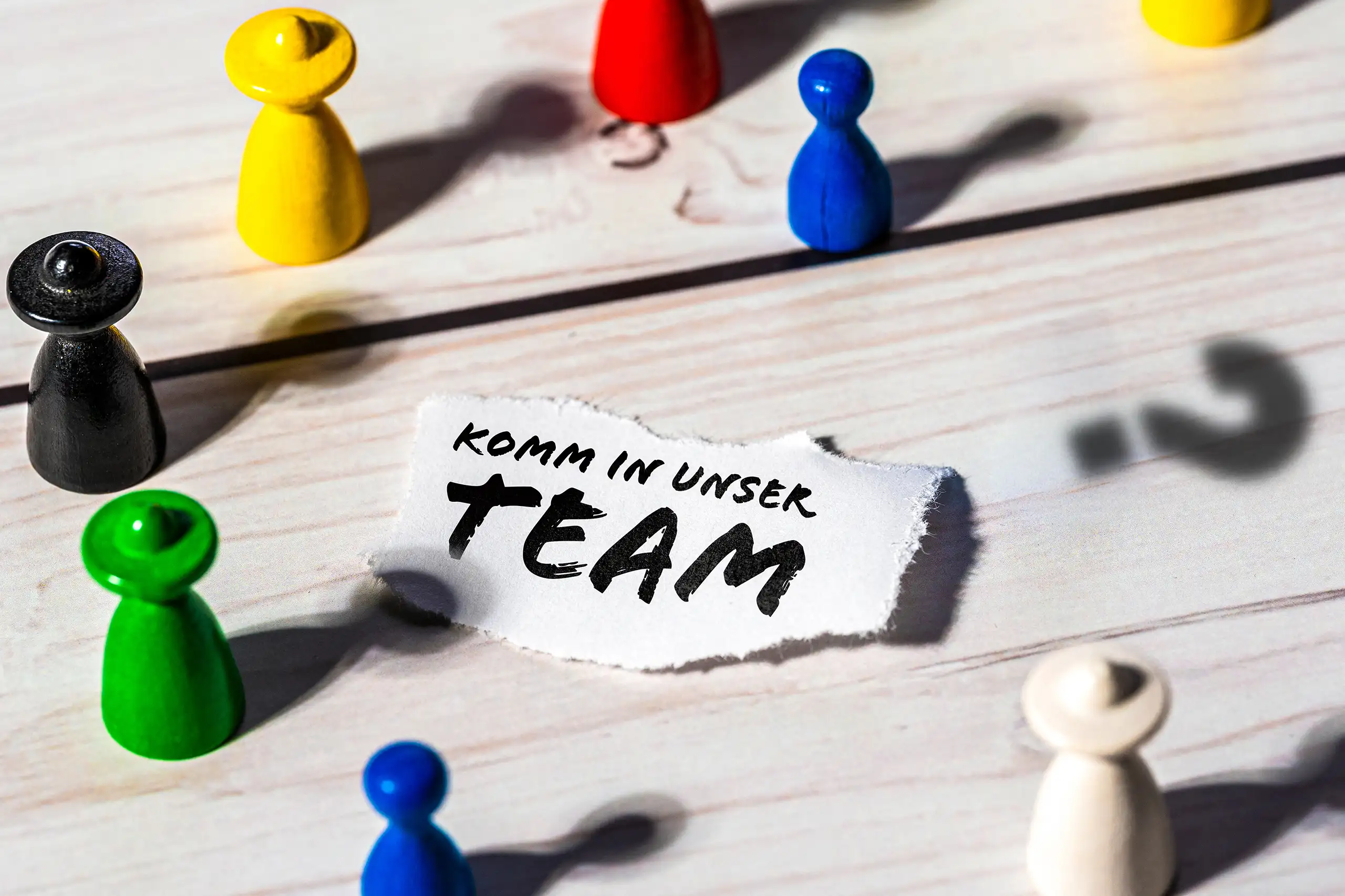 Bunte Spielfiguren auf einer Holzoberfläche umgeben einen Papierzettel mit der Aufschrift Komm in unser Team, daneben ist der Schatten eines Fragezeichens zu sehen.