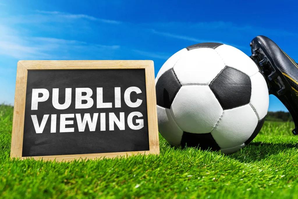 Public-Viewing-Schild neben Fußball und Fußballschuh auf grünem Rasen.