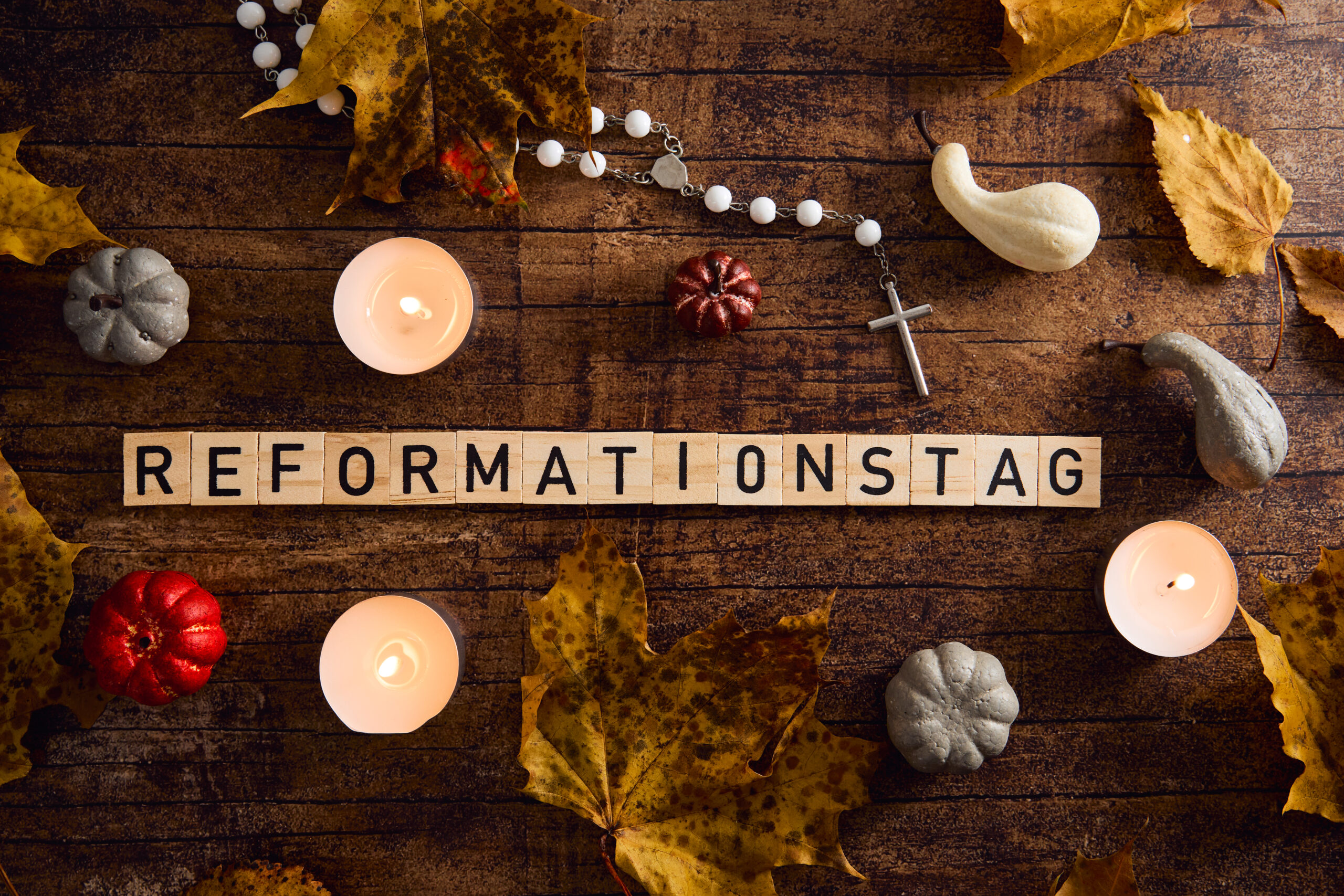 holzbuchstaben bilden das wort reformationstag auf holztisch, umgeben von kerzen, kreuz, herbstlaub und kleinen zierkürbissen.
