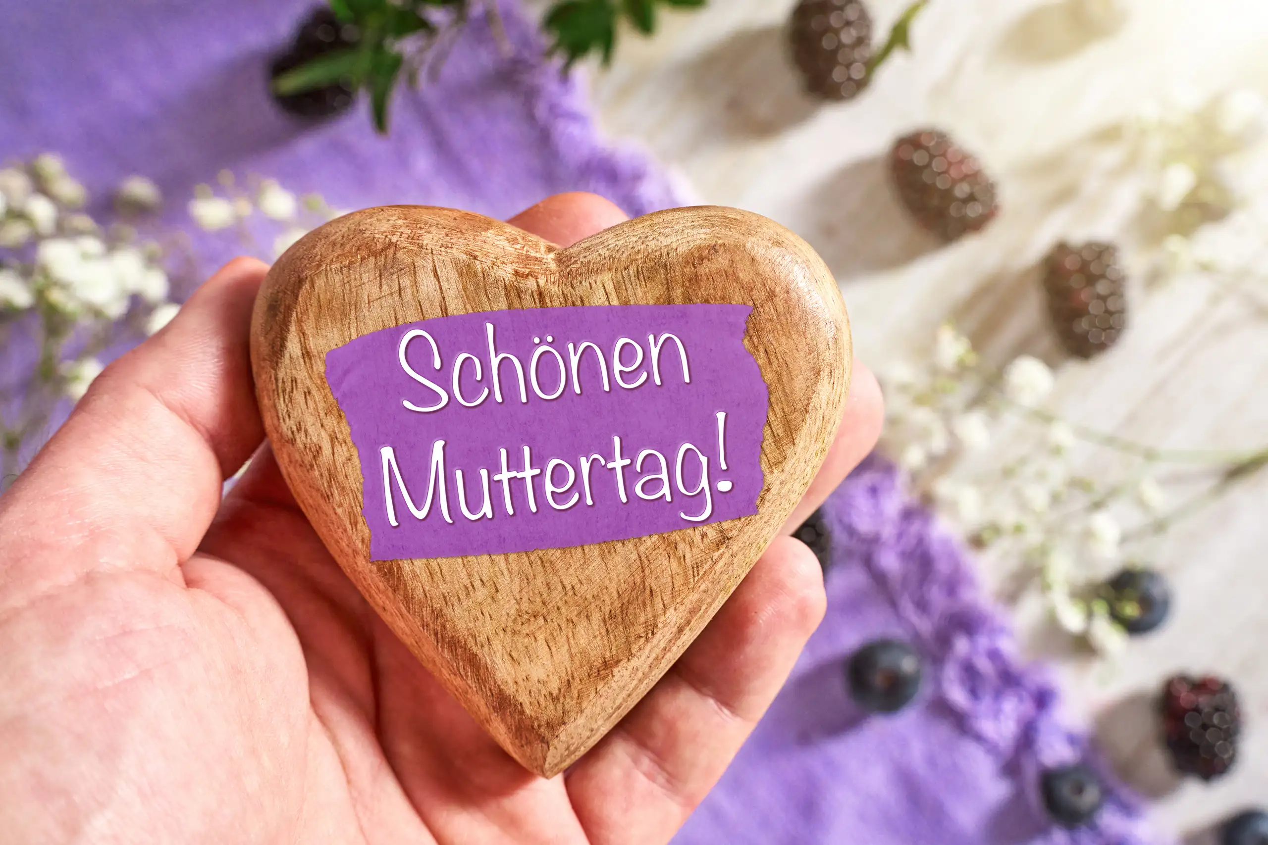 SCHOENEN MUTTERTAG MUTTERTAGSGRUSS BILDER schönen muttertag muttertagsgruß auf holzherz in einer hand mit violettem hintergrund, blumen und dekorativen frühlingsdetails.