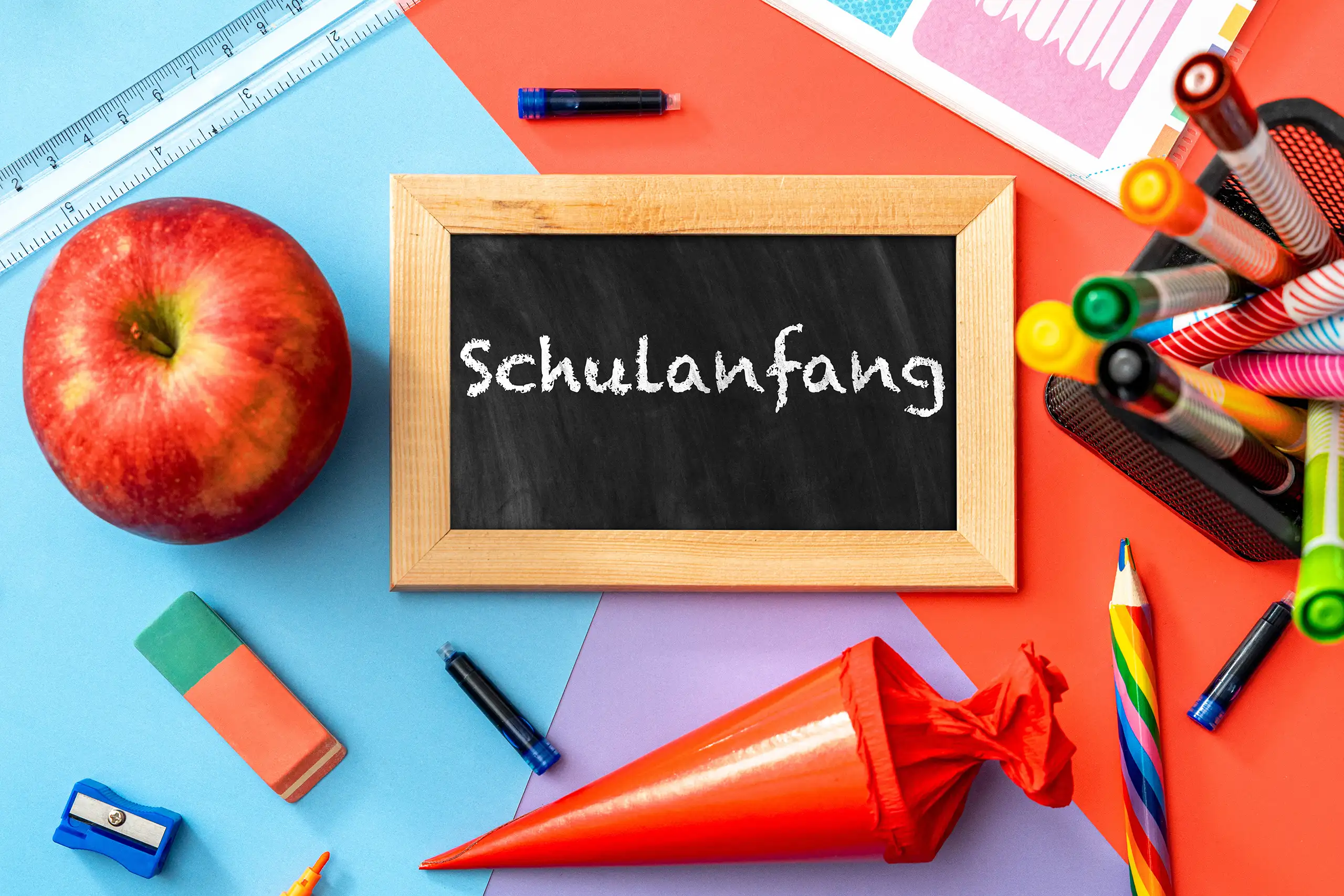 Schulanfang Bild mit schwarzer Schultafel, roter Schultüte, Apfel und bunten Schulmaterialien zum Schulstart