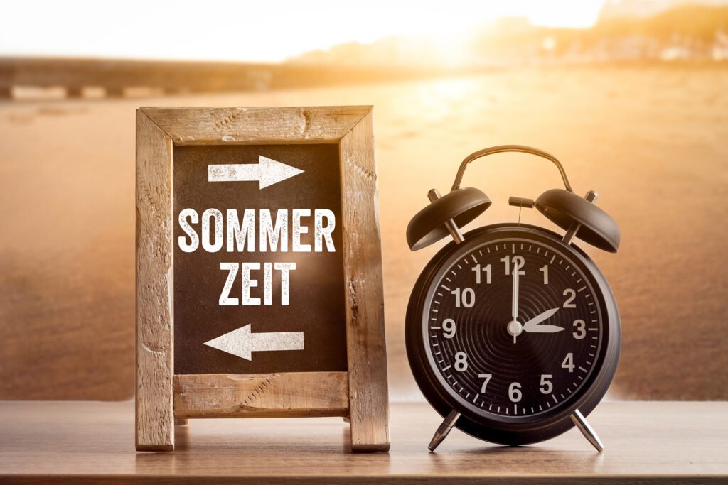 Sommerzeit, Schriftzug auf einer Tafel neben einem Wecker am Strand