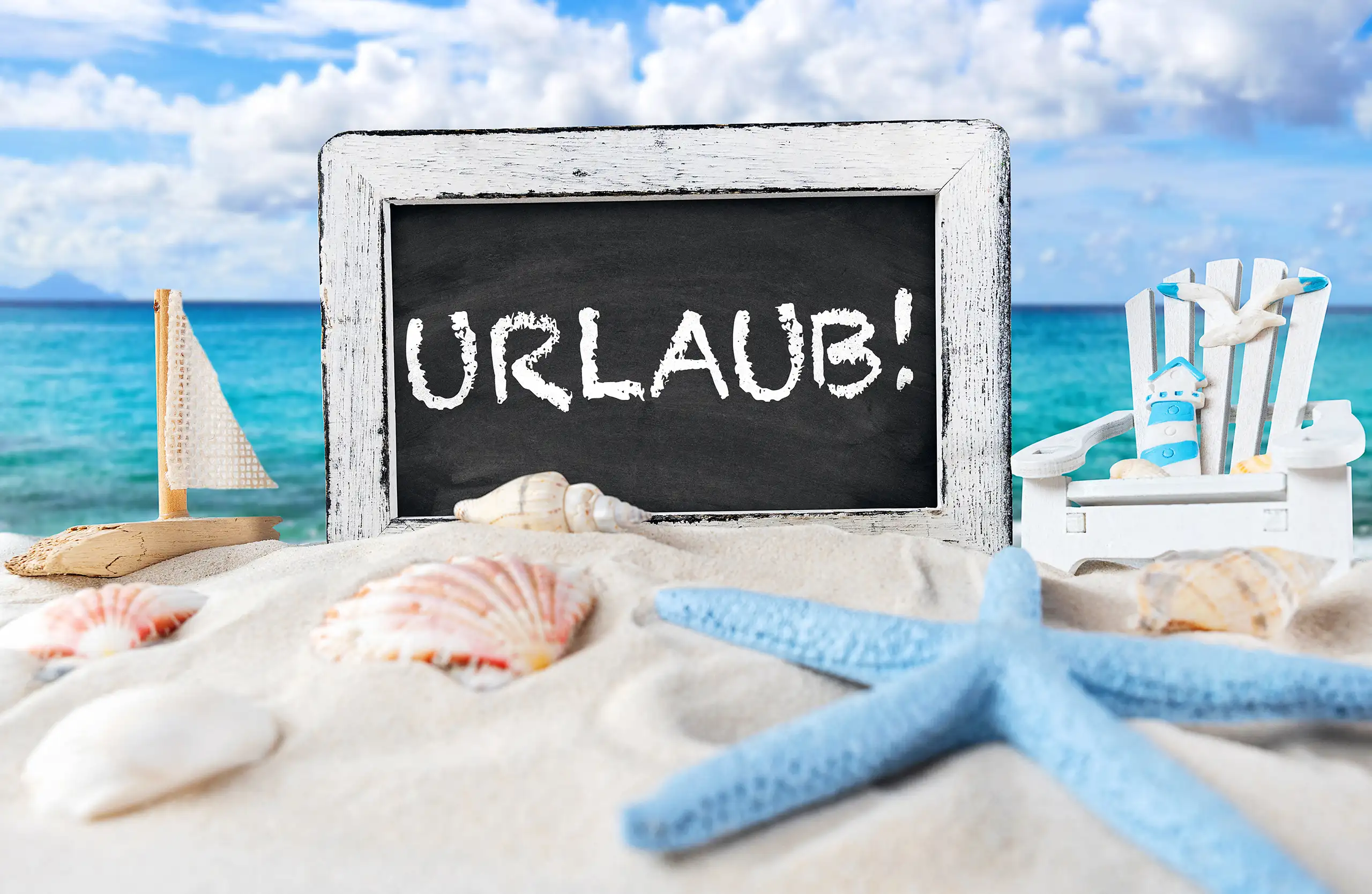 Urlaub Schriftzug auf schwarzer Kreidetafel im Sand vor blauem Meer