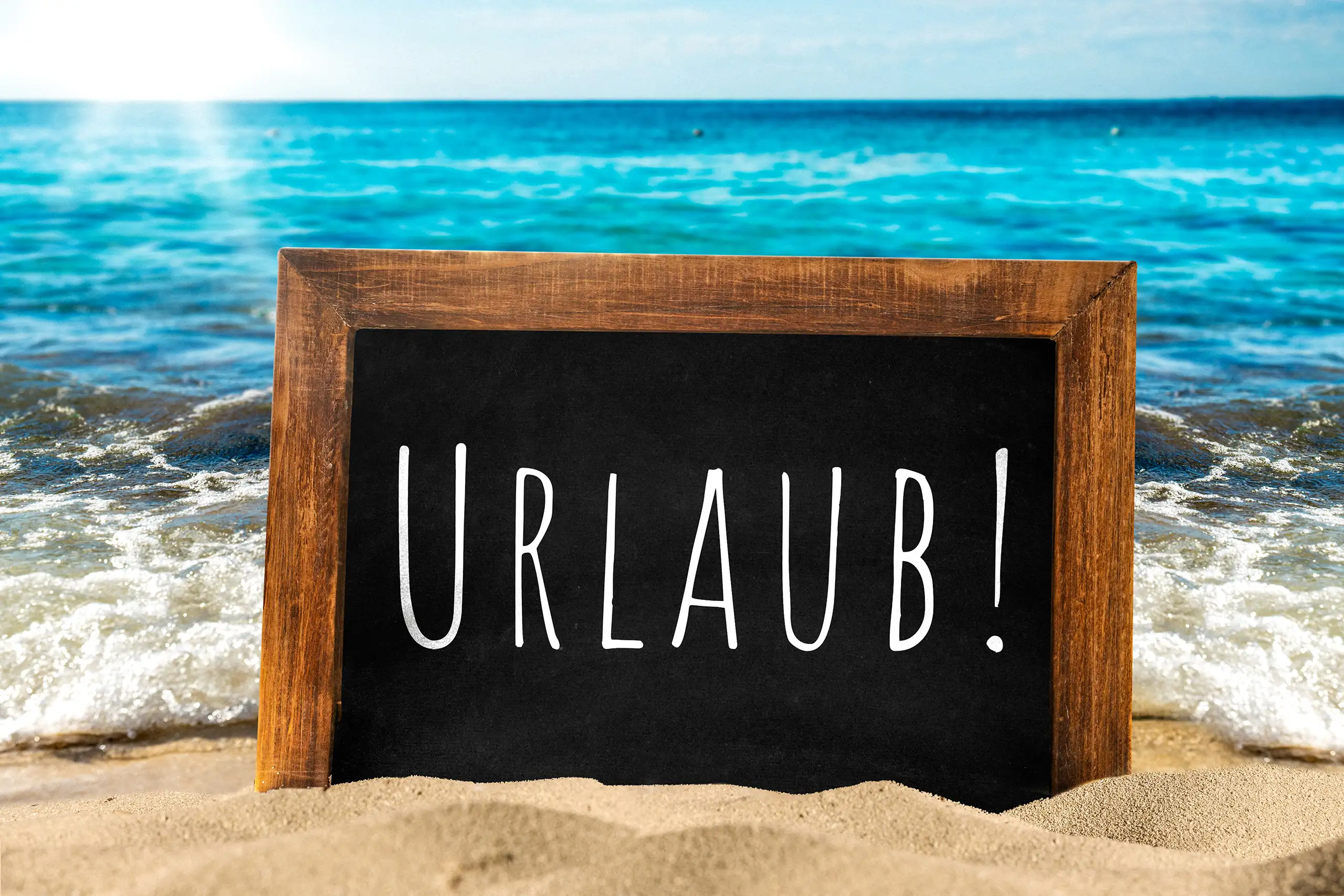 Urlaub Bild mit schwarzer Kreidetafel und Schriftzug Urlaub im Sand vor blauem Meer