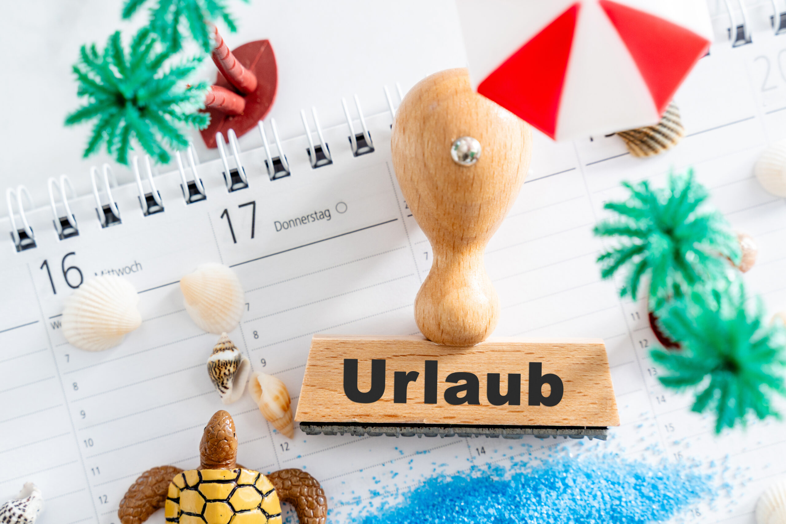 URLAUB BILDER HOLZSTEMPEL MIT DER AUFSCHRIFT URLAUB AUF EINEM KALENDER, UMGEBEN VON MUSCHELN, EINER MINI-PALME UND BLAUER DEKO holzstempel mit der aufschrift urlaub auf einem kalender, umgeben von muscheln, einer mini-palme und blauer deko